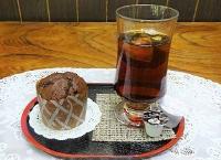 白いテーブルの上に枠が茶色で中が黒のお盆に乗ったカップケーキとグラスに入ったコーヒーの写真
