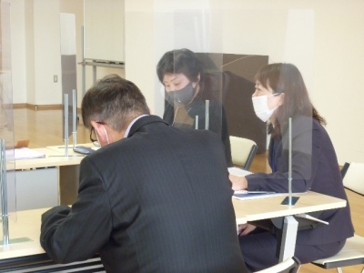 セパレータで仕切られたテーブルの上で、マスクを着けたスーツ姿の3人が話し合っている写真