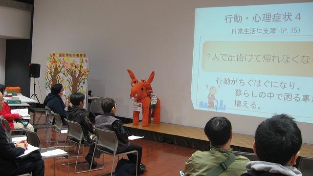 壇上に置かれた認知症サポーターキャラバンのマスコットキャラクターロバ隊長と着席して白いスクリーンに投影された行動・心理症状に関する説明イラストを見つめる認知症サポーター養成講座に参加した男女たちの写真