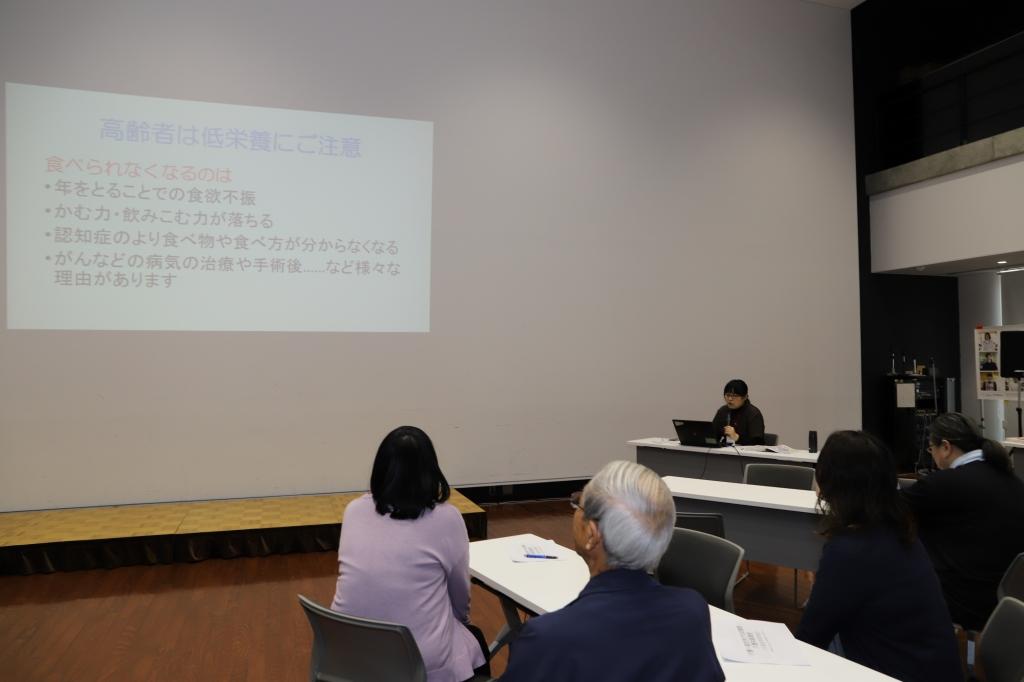 白いスクリーンに投影された高齢者の栄養摂取に関するメッセージについて説明をする女性とそれを見つめる参加者の男女4人の写真