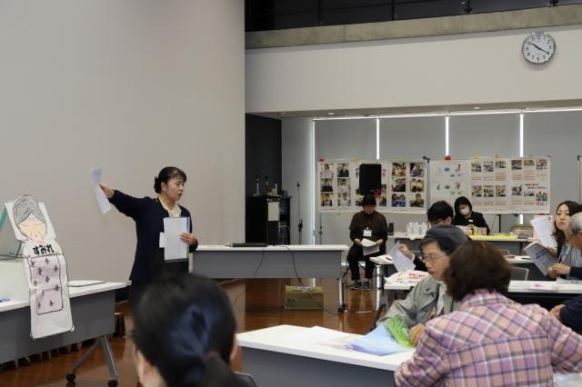 すみれと書かれた高齢女性の工作イラストを前に説明をする女性と説明を聞きながらパンフレットに目を通す参加者の男女の写真