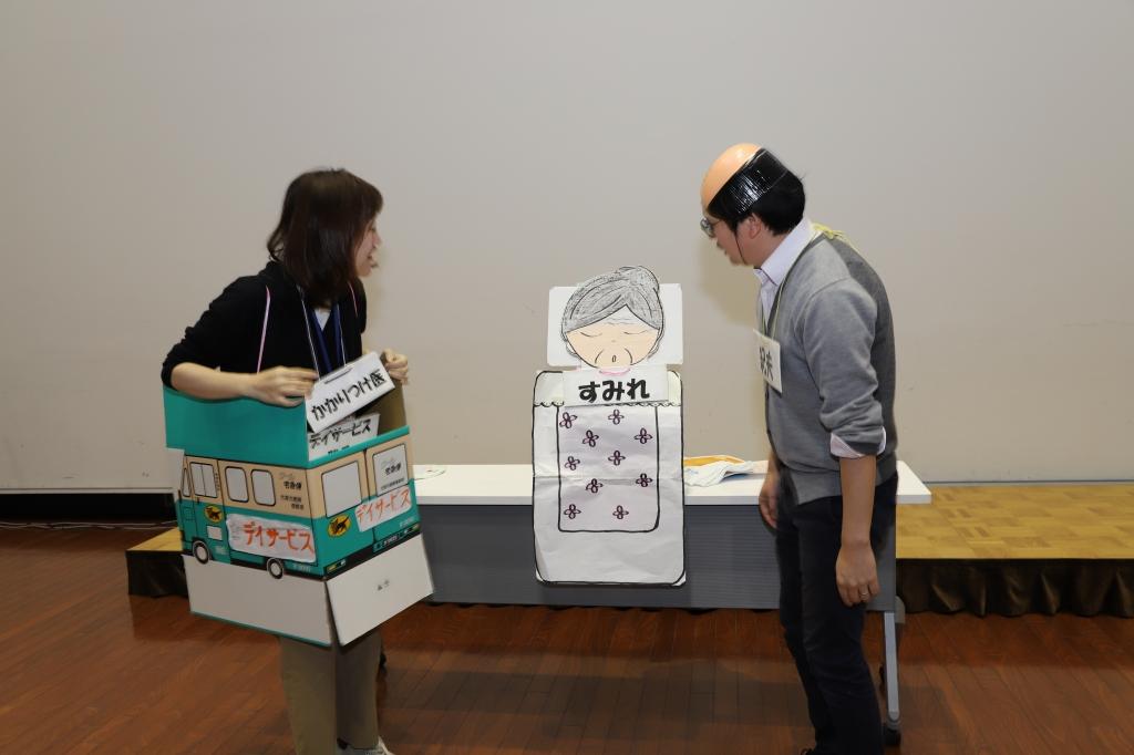 紀夫と書かれた札を首に下げた禿頭のかつら姿の人物とデイサービスと書かれたダンボールを被りかかりつけ医と書かれた札を首に下げた女性がすみれと書かれた高齢女性の工作イラストに問いかけている寸劇の様子の写真