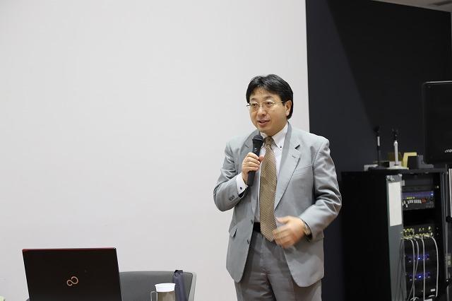 ミニセミナー会場で立ったままマイクを持ち話をする眼鏡をかけたスーツ姿の男性の写真