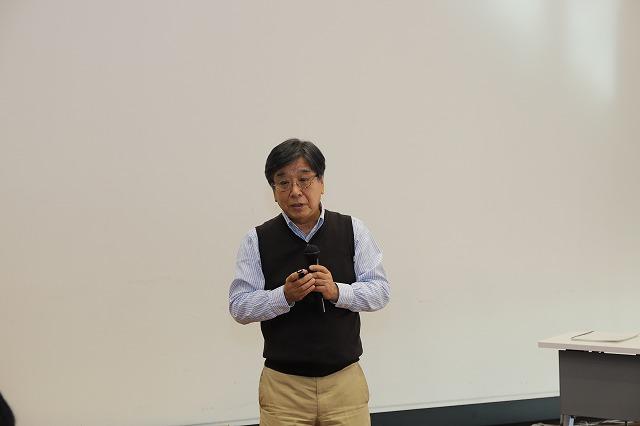 白い壁を背景に立ったまま両手でマイクを持つ眼鏡をかけた男性の写真