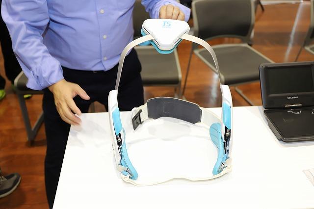 白い机に置かれた白と水色を基調とする体幹訓練器具を手にする青い長袖シャツを着た人物の写真
