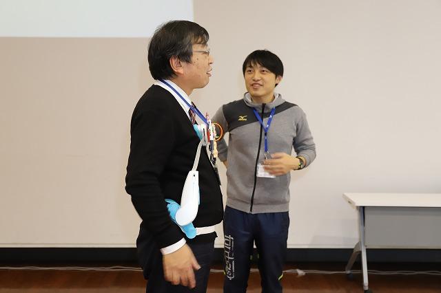 体幹訓練器具を装着したまま立っている眼鏡をかけたスーツ姿の男性とその様子を見つめているグレーのパーカーを着た若い男性の写真