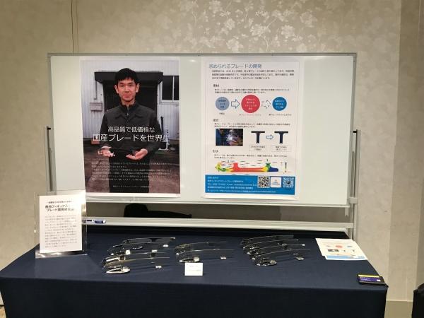 燕市フィギュアスケートブレードの概要を説明したチラシとブレードのつくりを表したチラシの2枚が貼られているホワイトボードと、展示用ブレードが置かれている写真