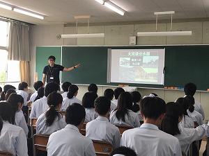 スクリーンに表示した写真を説明している黒いシャツの講師と、座って聞いている生徒たちの写真