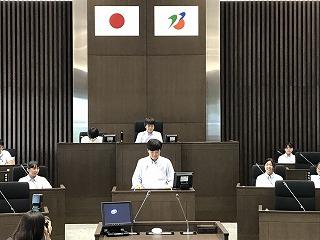 議場で、前方部分で立ち上がっている男子生徒と、周りで着席している生徒たちの写真