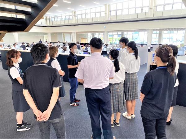 明るいオフィスで、説明をする若い社員と、話を聞いている生徒と関係者の写真
