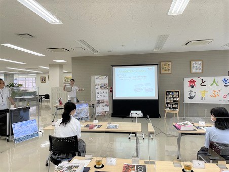 社員がスクリーンでプレゼンをしていて、その最中に商品を手に持ち説明しているところを女子生徒2人が聞いている写真。
