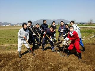 作業着を着た男女が畑の前でスコップを手に持ち記念撮影をしている写真