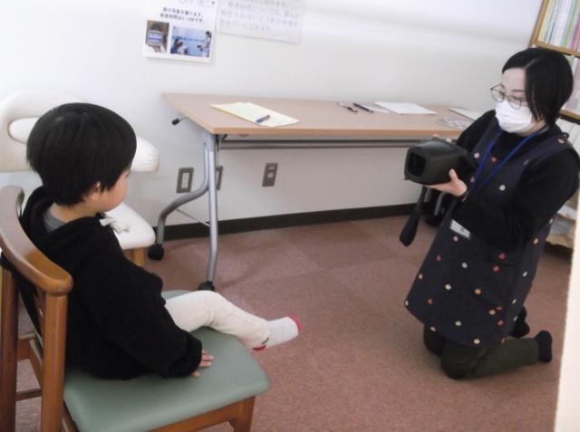 椅子に座っている子供と、膝立ちで子供を撮影している女性の写真