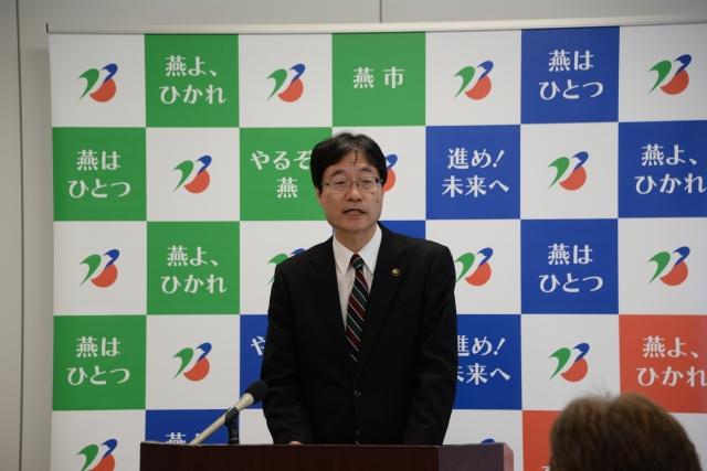 燕市のポスターを背に話しているスーツ姿の男性の写真