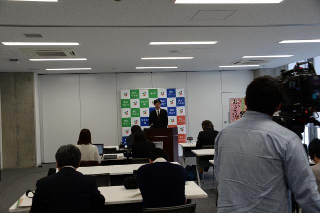 燕市のポスターを背に話している男性と、座って聞いているスーツ姿の人々と、それらを撮影しているカメラマンの写真
