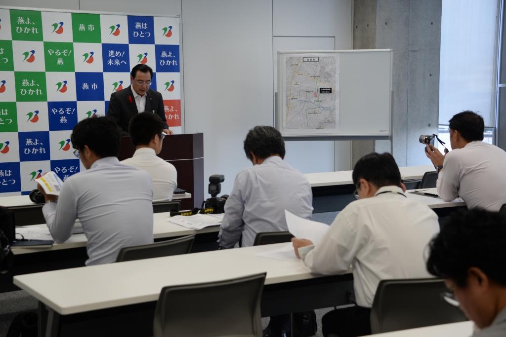燕市のポスターを背に発表をしている教育長の男性と、座って聞いている関係者たちの写真