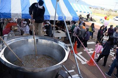 巨大な釜で食材を混ぜている男性と、後ろの屋台に集まっている人たちの写真