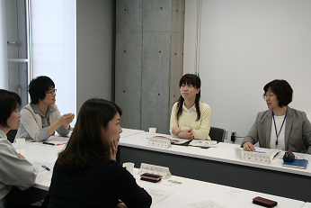長方形に並べられた資料が置かれた机の手前の椅子に座りながら話し合う5人の女性たちの写真