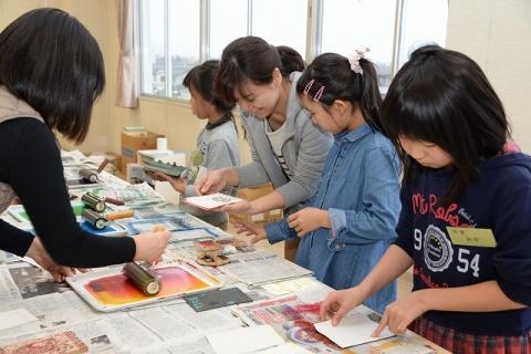 完成した版画の作品を見て笑顔を浮かべる女性と女性のそばにいる子どもやローラーにインクを塗り込む女性などそれぞれ版画作りに取り組む様子の写真