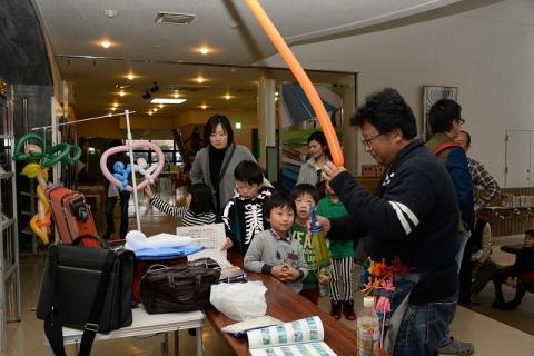色とりどりのバルーンアートが3つ飾られたブース内でオレンジ色の細いゴム風船と空気入れを手にする男性とブースに来た子供や女性たちの写真