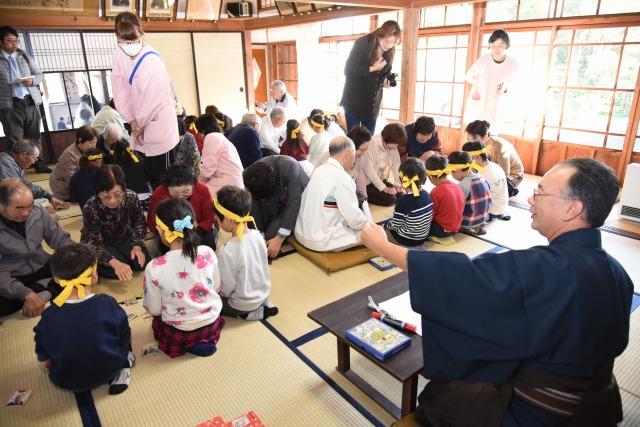 かるたの読み札を読む男性と、真剣に聞き入っている参加者たちの写真