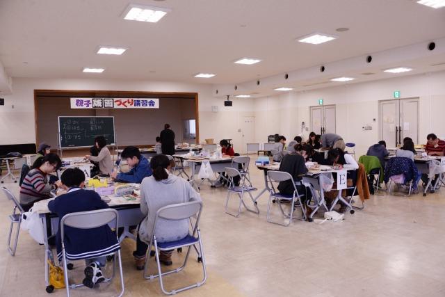 「親子版画づくり講習会」と書かれた垂れ幕がある部屋で、グループに別れて机を囲んで作業をしている参加者の写真