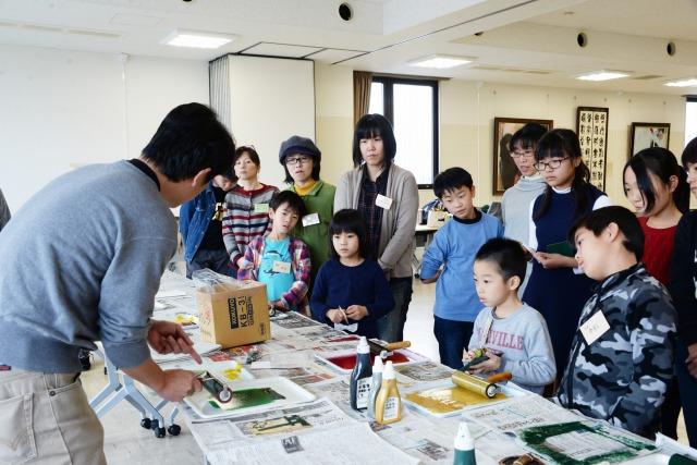 新聞紙が敷かれた広い机で、ローラーを片手に説明をする男性と、机を挟んで聞いている子どもたちの写真