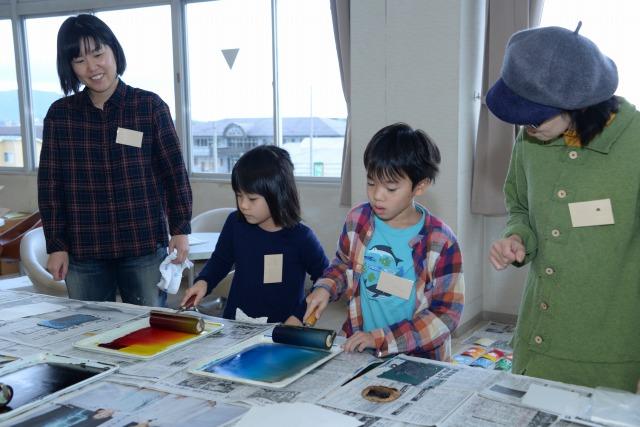 赤と黄色、青などの色がついたローラーを動かしている、少女と少年とその脇で笑顔で見守る二人の女性の写真