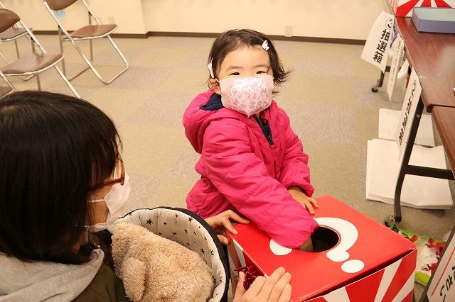 抽選箱に手を入れてこちらに笑顔を向けるピンクのコートを着た少女の写真