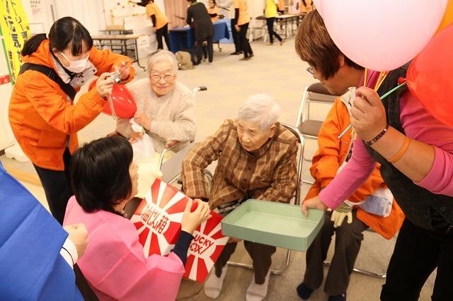 椅子に座りながら抽選箱に手を入れている女性と抽選箱を低くして持っている女性とを写真に収めているスタッフの写真