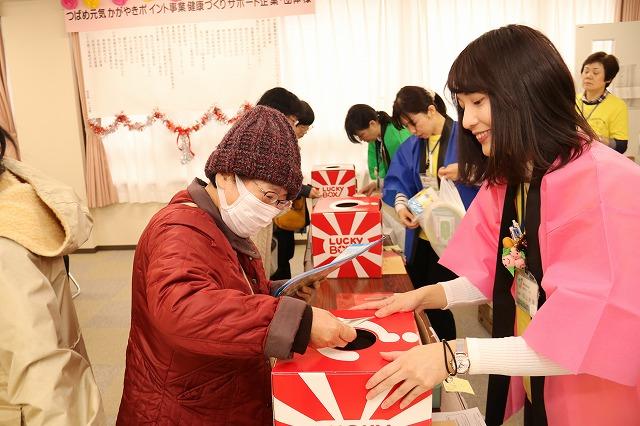 抽選箱に手を入れているニット帽の女性と箱を抑えているピンクのハッピの女性の写真
