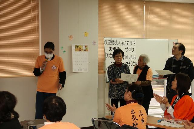 左側にマイクを持つ男性、ホワイトボードの前に立つ女性2名と右横に立つ男性1名が、紙を手に持って司会を進行してる様子の写真