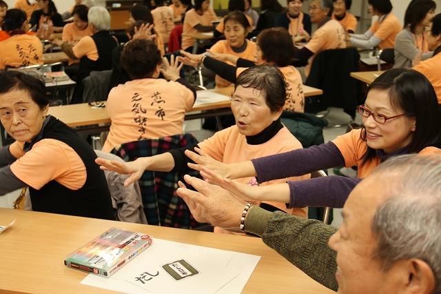 おそろいの薄いオレンジのTシャツを着た年配の方々が、両手を前に出し楽しそうにプログラムに参加している写真
