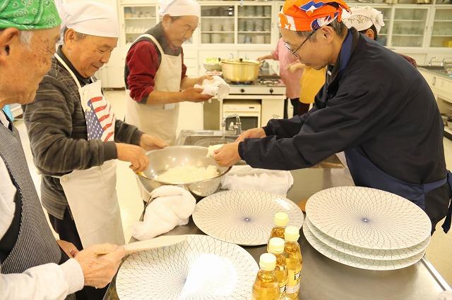 お米の入ったボウルや調味料とお皿が置かれた調理台を囲み、エプロンとバンダナをつけた男性数名が手にしゃもじを持って料理の準備をしている写真