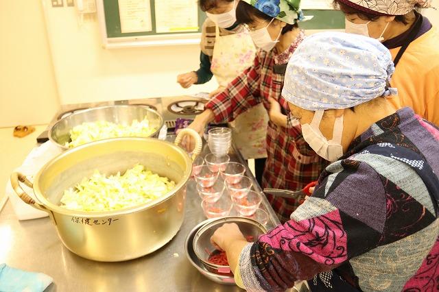 大きな鍋と盛り付け用の器が並んだ調理台の前で、バンダナとマスクとエプロンをつけた女性4名が料理の準備をしている写真