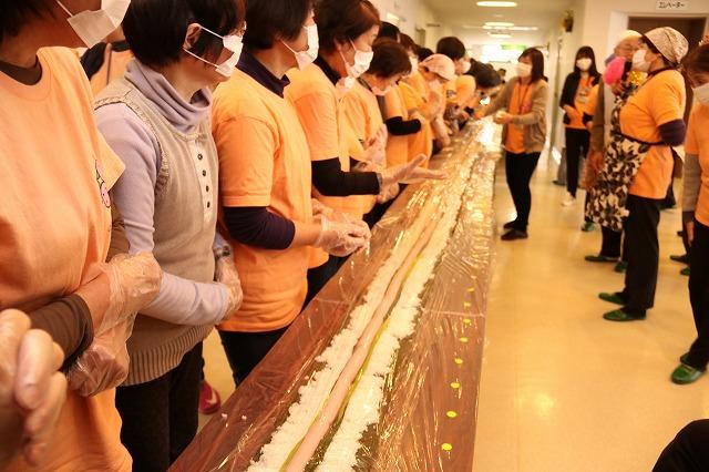具材が並べられた長いのり巻きが置かれた長いテーブルを前に、横一列に並んで待機している参加者の写真