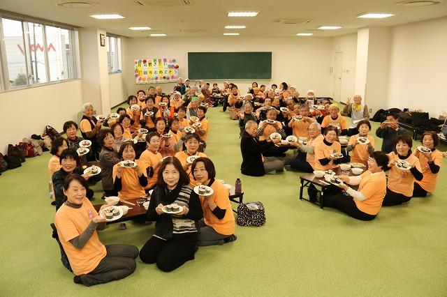 室内に複数置かれたテーブルを囲んで座り、出来上がったのり巻きを掲げている参加者全員の記念写真