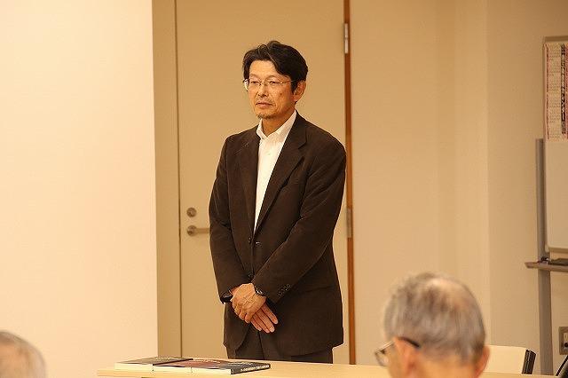 参加者と向かい合わせの机の前に立つ、茶色い上着の男性の写真