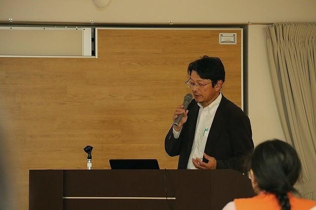 マイクを手に演題で話をしている、茶色い上着の男性の写真