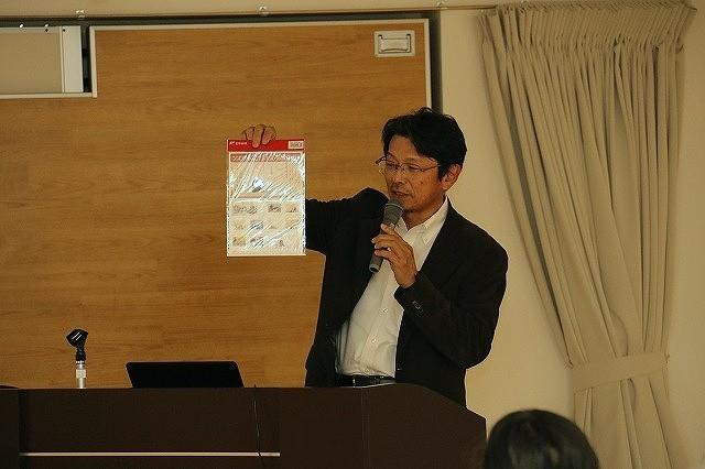 演題で、右手に資料を持ち上げて見せながらマイクを手に話しをしている茶色い上着の男性の写真