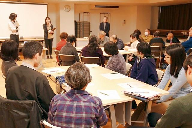 7人がけの複数の机に座った大勢の人々の前で司会をしている二人の女性と、近くに立っている白髪の男性の写真