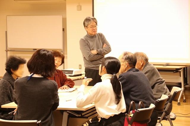7人がけの机に座った1つのグループの人々の横に腕組みをして立っている白髪の男性の写真