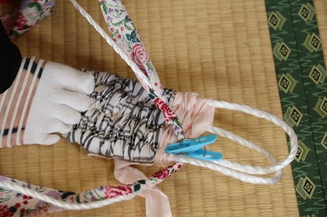足で押さえながら鼻緒用の紐に赤い花の模様の布を巻き付けている様子の写真