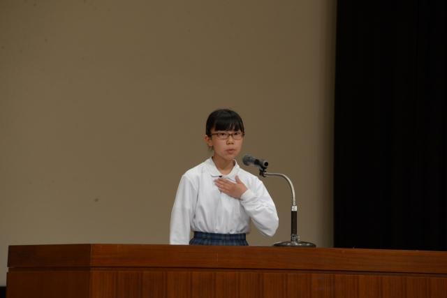 壇上で左手を胸にあてながらスピーチをしているメガネをかけた女子の写真