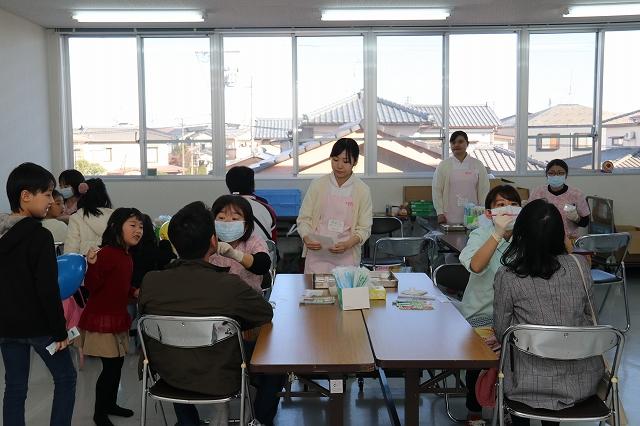 医療器具を持ちパイプ椅子に腰かけ男性と女性それぞれの歯科受診を行う2人のマスク姿の女性とその様子を覗きこむ青い風船を持った女の子と白い冊子を右手で握っている子どもの写真