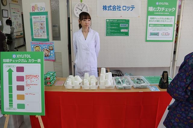 試供品のロッテ社のキシリトールガムや白い紙コップとメトロノームなどが置かれた長机を前に立っている白衣姿の女性と長机の前にあるガムをよく噛んでいるかどうかの問いに対しカラフルなシールによる回答が多数貼られているシール投票のボードの写真