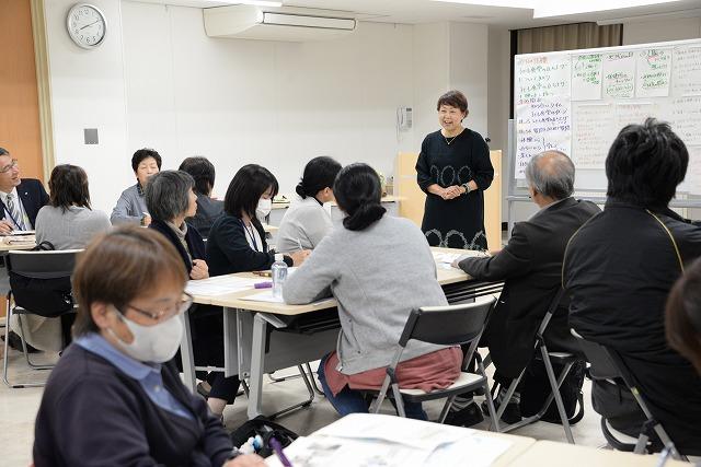 "セミナーの議題について書かれた用紙が貼られているホワイトボードのそばで両手を重ね合わせて話をする黒い服を着た女性とテーブルに腰かけ女性の方を向いて話を聞いている男女の参加者たちの写真 "