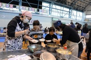 テーブルの上のボウルに入っている汁物をおたまで皿によそう2人の女性と皿を受け取る女子生徒2人の写真