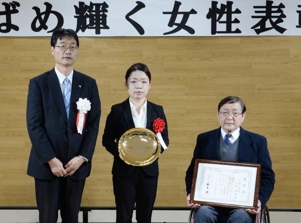 市長と並んで立つ盾を持つ女性と賞状を持つ車椅子の男性の写真