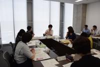 壁側の大きな窓の手前に四角に並べられた机の周りに座り資料を見ながら会議に参加する女性たちの写真